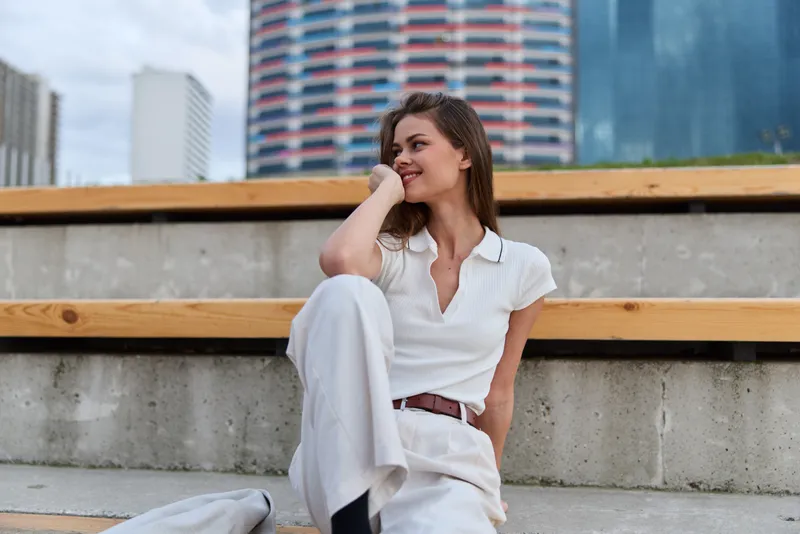 Young woman in white casual outfit sitting relaxed outdoor in urban environment with modern buildings smiling