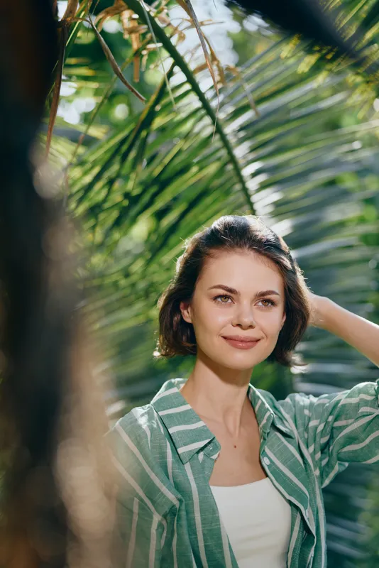 Portrait of a smiling woman standing among lush tropical plants, wearing a light striped green jacket and white top, bathed in soft natural light.