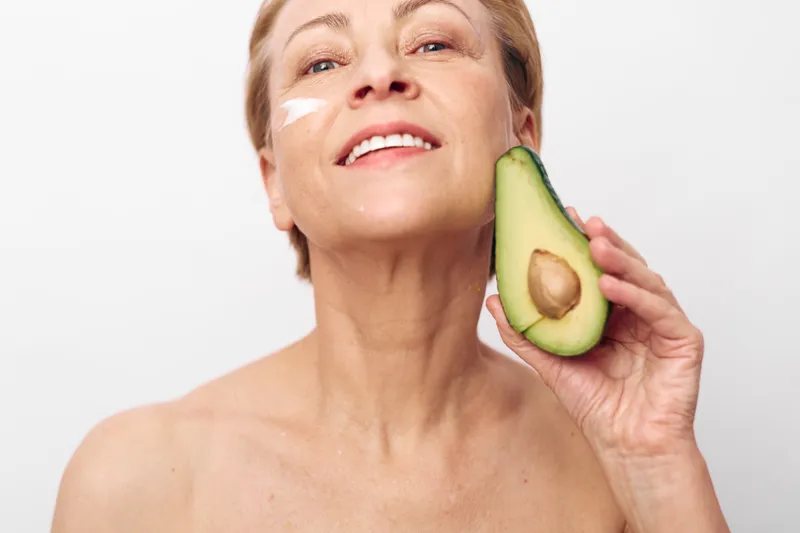 Mature woman with glowing skin holds fresh avocado near face, applying cream on cheek. Natural skincare and healthy lifestyle concept with bright soft lighting and minimal background.