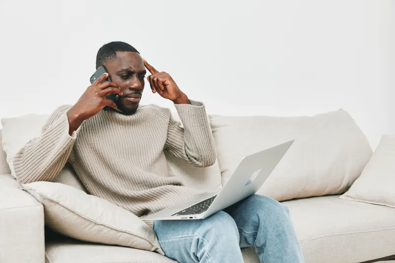 Young Black man in casual sweater expressing frustration while talking on the phone and working on a laptop at home