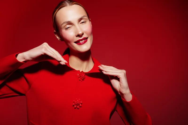 Smiling woman in red sweater poses with hands near face on red background. Studio portrait with bright lighting and joyful expression. Concept of fashion and happiness.