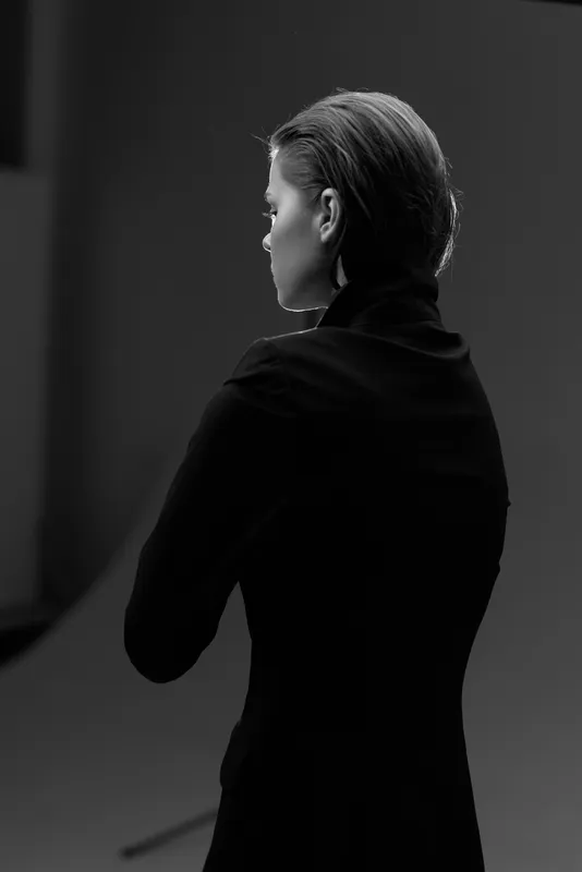 Black and white portrait of a serious woman in black clothing with crossed arms standing in a dark studio.
