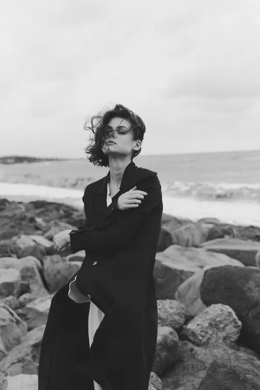 Fashion model in a long coat and sunglasses on a windy coast, rocks and sea behind, creating a moody monochrome scene with wind swept hair and bold urban attitude.