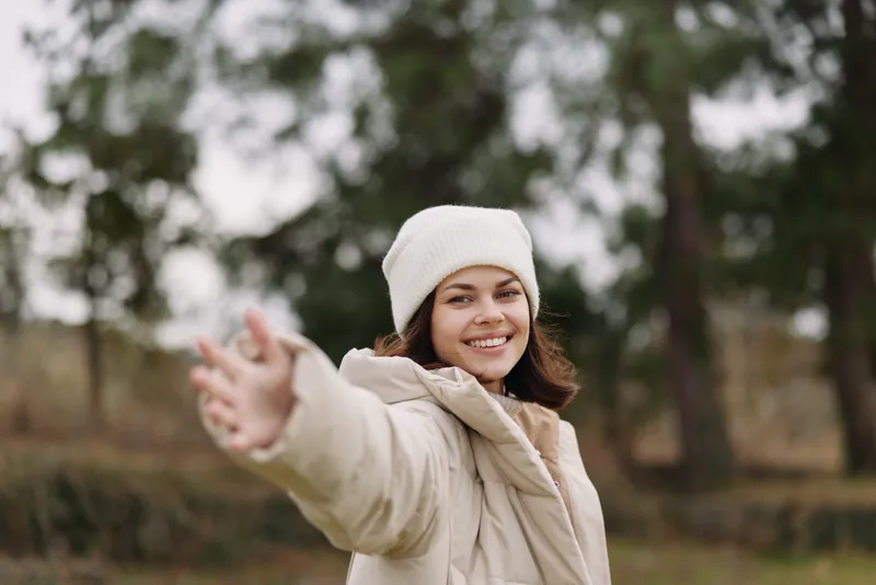 Woman in a warm beige coat and white beanie smiles warmly outdoors, extending her hand in a friendly gesture against a blurred park background.