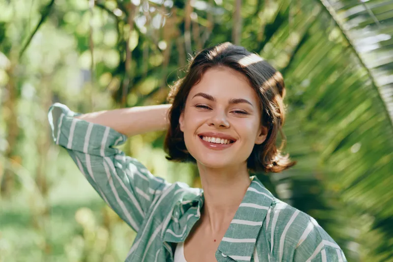 A cheerful woman smiles softly as she enjoys a sunny day outdoors. The portrait captures her relaxed pose amid lush greenery and natural light, conveying warm mood and confidence.
