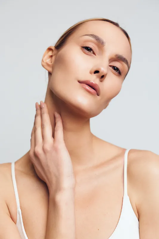 A young woman with clear skin poses in a studio, touching her neck with a soft expression and a white tank top, captured under studio lighting for a clean beauty moment.