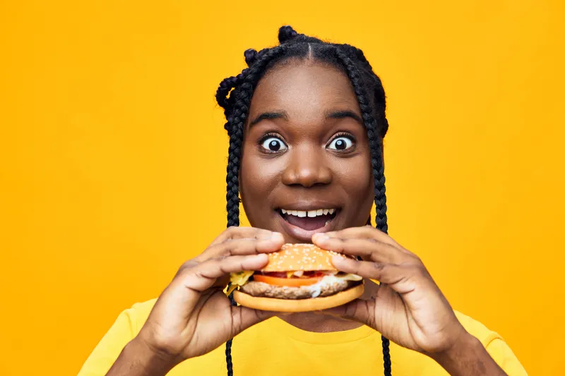 Happy young Black woman holding a delicious burger against a vibrant yellow background, showcasing joy and excitement about healthy eating