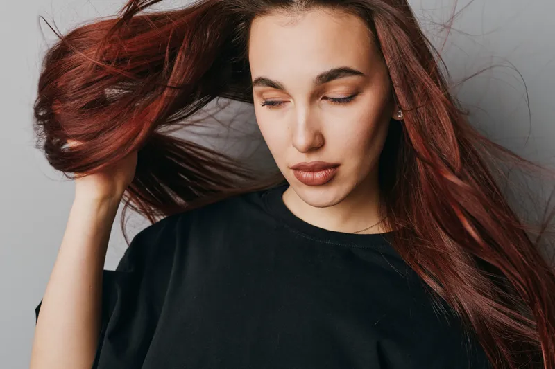Woman with long red hair and closed eyes wearing black sweater posing on gray background. Female model with natural makeup and smooth skin showing hair movement in studio.