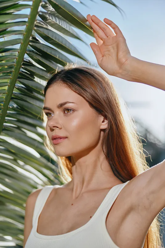 young woman shielding her eyes from the sun with hand near tropical palm leaves in bright outdoor natural light, casual white tank top, serene expression
