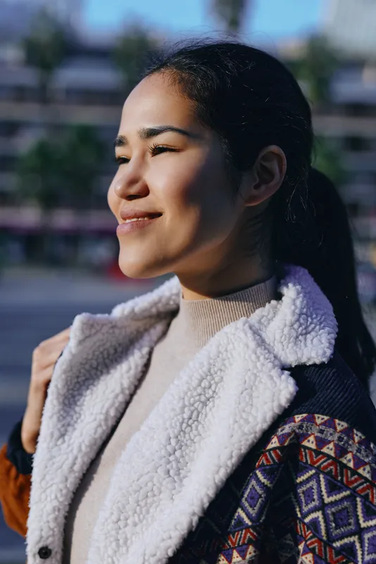Smiling, sunlit subject wearing shearling coat and patterned jacket outdoors, warm light, wind, street setting, casual fashion moment captured in natural glow.