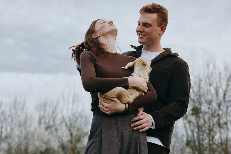 A young woman joyfully holds a baby goat while a young man stands behind her smiling in a casual outdoor setting with bare trees in the background.