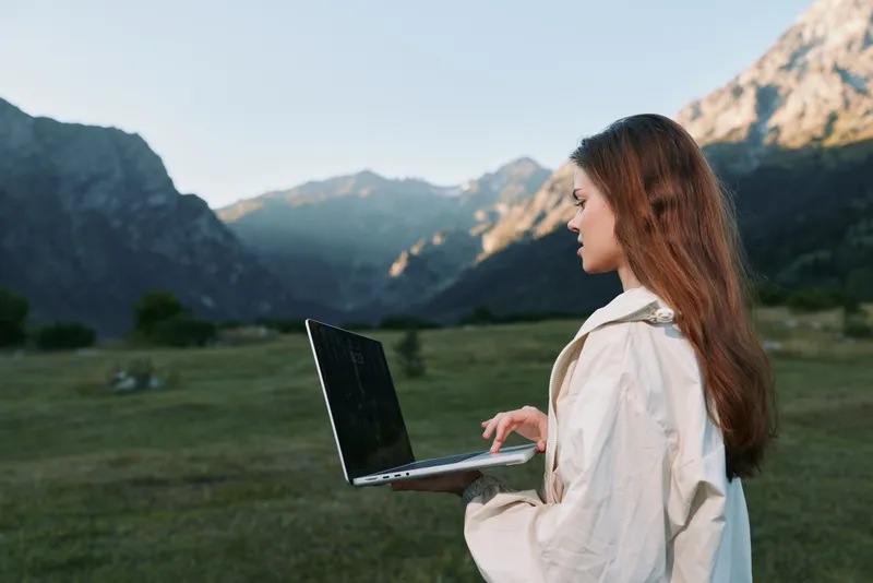 A focused professional woman stands outdoors with a laptop in a scenic valley, typing thoughtfully as daylight highlights her calm, determined demeanor and productive mood for a dynamic work scene.