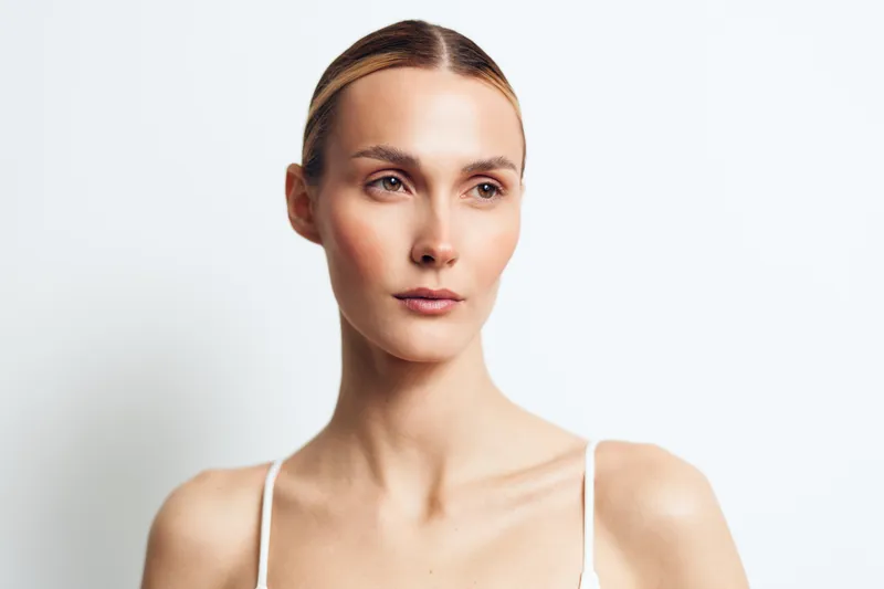 A young woman with clear skin and natural makeup looks thoughtfully to the side against a white background. Concept for beauty, skincare, and natural lifestyle.