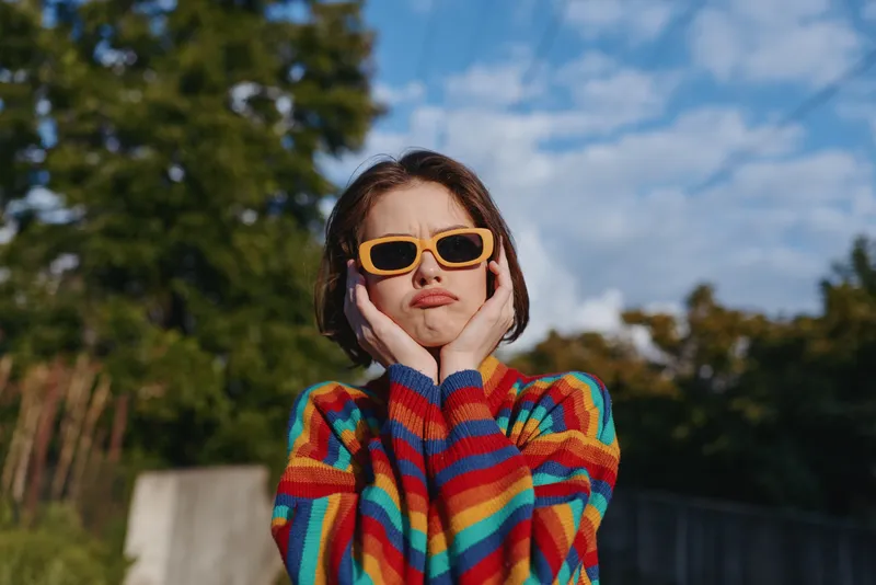 Young woman in rainbow sweater and yellow sunglasses posing outdoors with hands on face and playful pout, short brown hair, sunny park background for lifestyle and fashion.