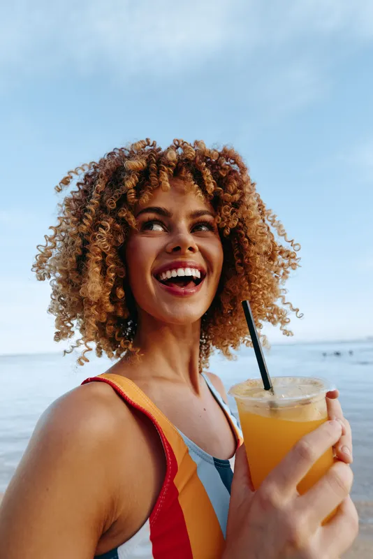 A young woman with curly hair smiles joyfully while holding a glass of orange juice at the beach under a clear blue sky during a sunny day.
