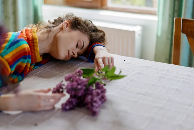 Young woman resting on a table indoors, colorful striped sweater, soft natural light, relaxed home moment, quiet table scene, lilac bouquet in foreground, peaceful mood