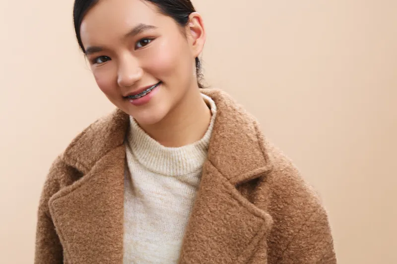 Asian woman wearing a warm brown coat smiles softly in a studio setting, presenting a friendly, approachable mood with calm lighting and a clean, minimalist backdrop.