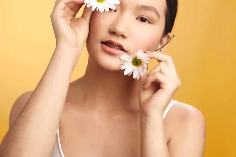 asian woman with delicate features smiles softly as she holds two daisies near her face, creating a fresh, natural portrait with a warm yellow backdrop.