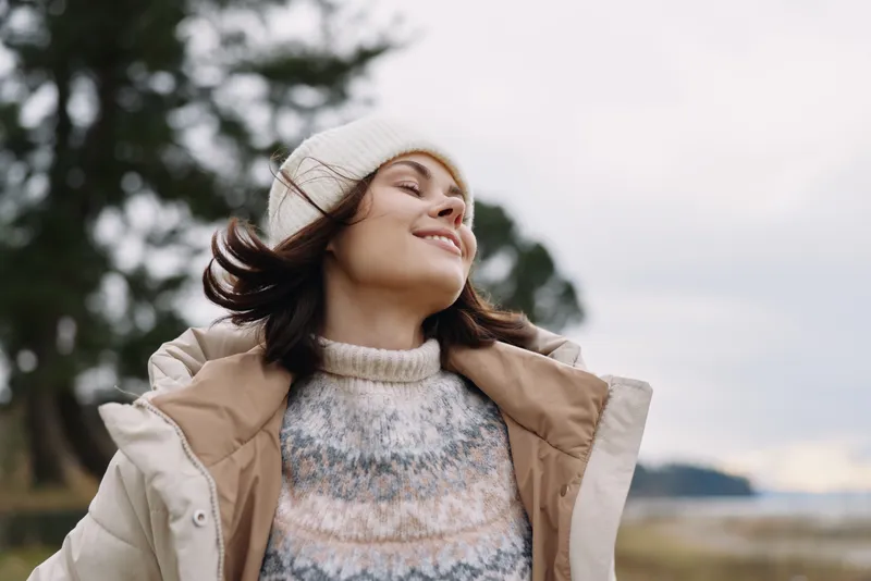 Woman outdoors, smiling as wind lifts her beanie and coat, wearing a cozy knit sweater in a natural setting with trees and open sky