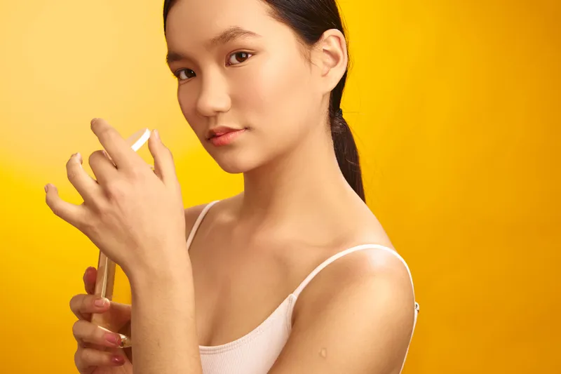 Asian woman portrait in close up, holding a makeup applicator near her lips, against a bright yellow backdrop, conveying beauty and focus in a clean studio setting.