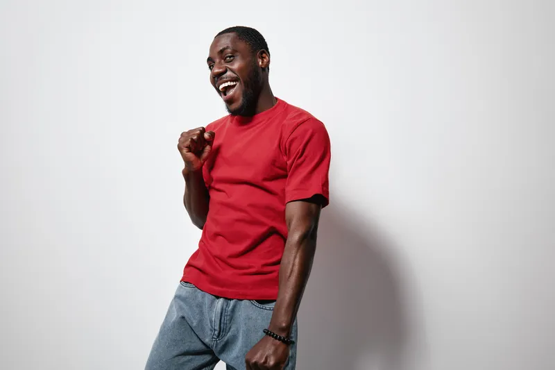 African American man with dark skin wearing red t shirt and jeans smiling and celebrating with fist pump against white background. Happy young male expressing joy and excitement.