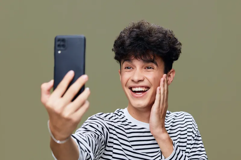Young man smiling while taking a selfie with a smartphone, wearing a striped shirt and showing joyful emotion on a simple olive background.