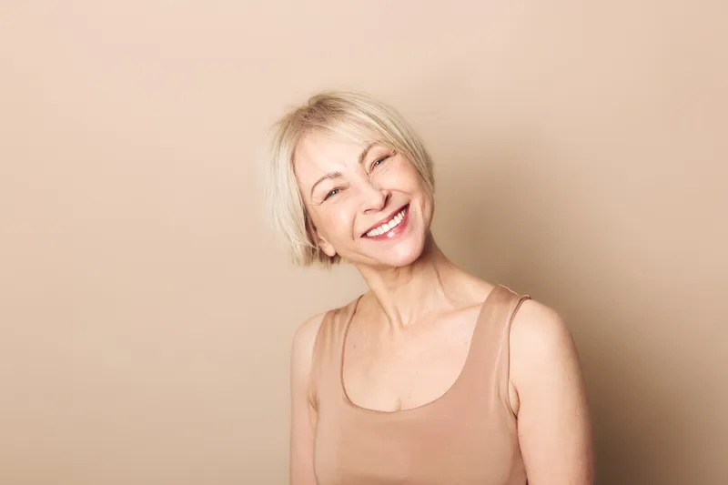 Smiling mature woman with short blonde hair wearing a sleeveless beige top poses against a neutral background. Concept of happiness, aging beauty, confidence, and natural smile.
