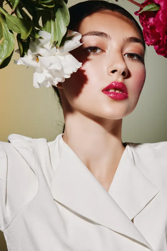 Close-up of a young woman with glossy pink lips and glowing skin wearing a white outfit. Flowers frame her face highlighting beauty, fashion, and creative glamour concepts.