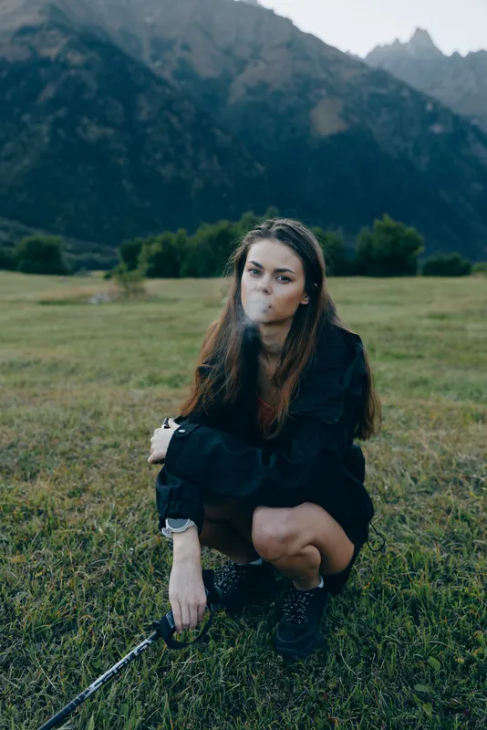 A young woman crouching in a grassy meadow with a distant mountain backdrop, smoking softly while looking thoughtful, dressed in dark streetwear, capturing a moody outdoor portrait.