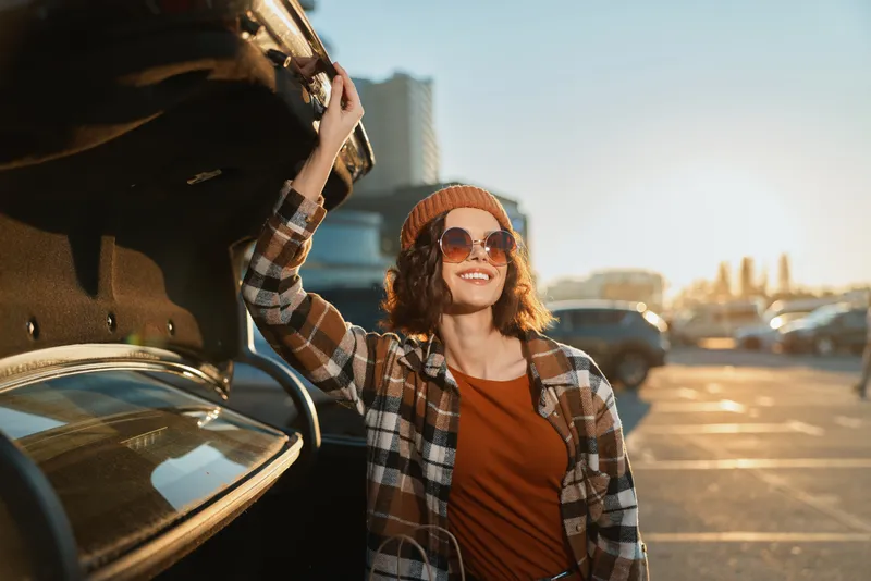 Woman at car trunk in parking lot smiling with sunglasses and beanie, urban portrait capturing authenticity and candid lifestyle, golden hour glow, mindful living and emotional storytelling.