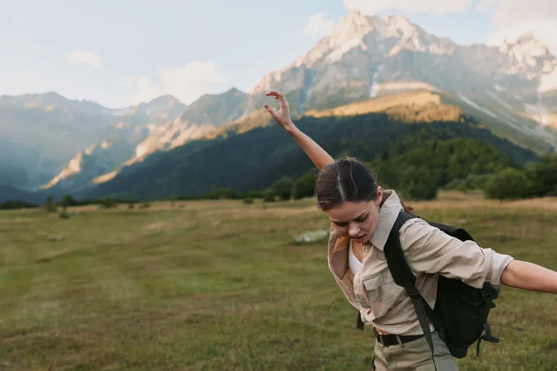 Young woman hikes in a meadow with mountains in the background, arms outstretched and joyful as golden light bathes the scene, light shirt and backpack ready for trek.