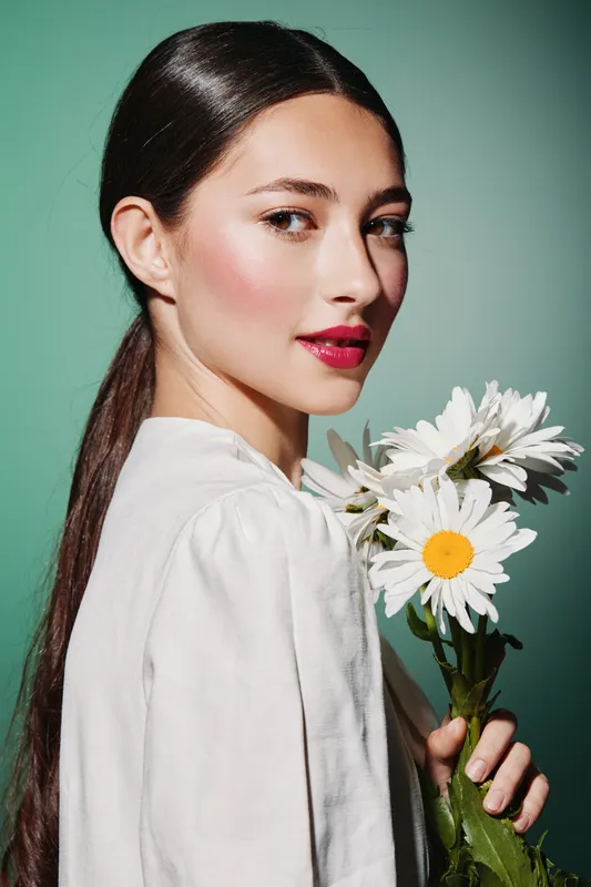 Young creative woman with sleek hair and vibrant red lipstick holding white daisies against a green background. Studio photo focused on beauty, fashion, and fresh style.