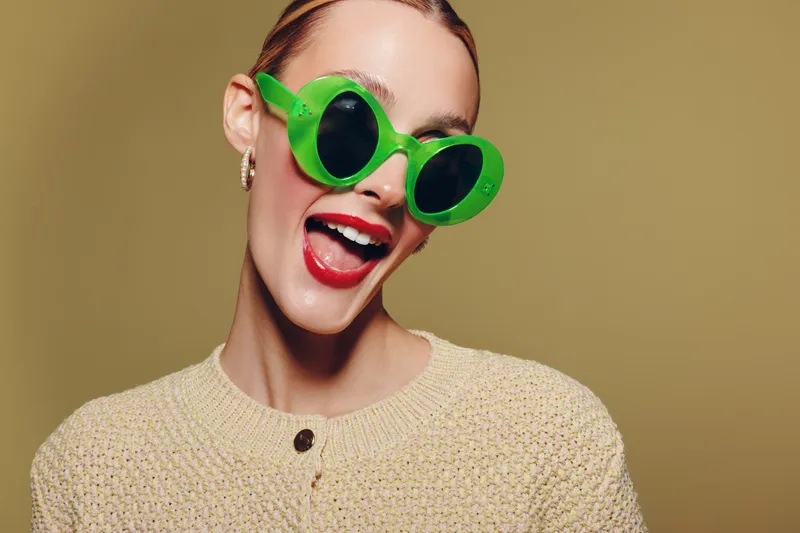 Happy young woman with bright red lipstick wearing green sunglasses and beige sweater smiles joyfully on yellow background. Concept of fashion, summer, and positive mood.
