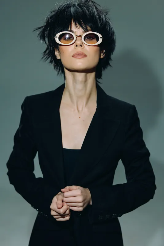 Stylish woman with short black hair wearing white round sunglasses and a black blazer posing confidently against a muted background.
