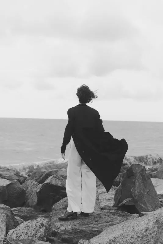 Woman stands on rugged rocks by the sea, a dark coat billowing in the wind, creating a stark silhouette against the vast horizon and a sense of mystery lingers in the chilled air.