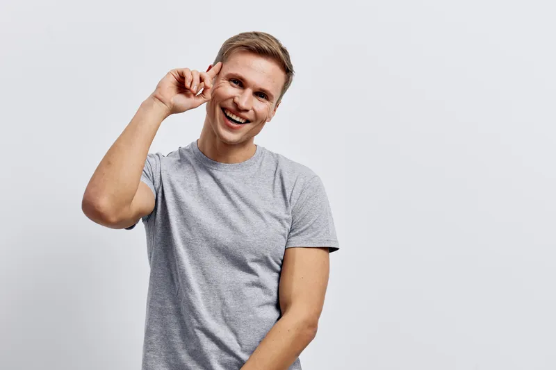 Smiling young man with short hair wearing casual gray t-shirt, looking cheerful while holding earphone near his ear, on plain white background. Lifestyle, happiness, casual, happiness concept.