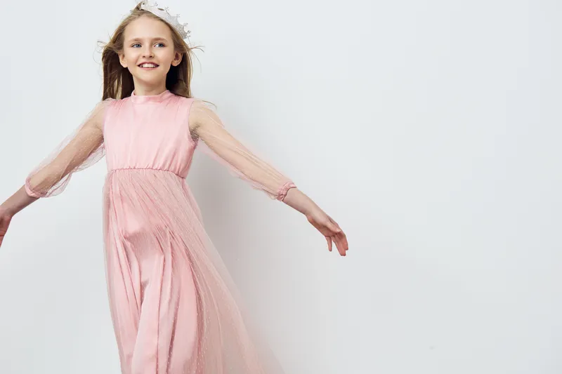 A joyful young girl in a flowing pink dress twirls gracefully, showcasing her radiant smile and innocence against a soft, light background