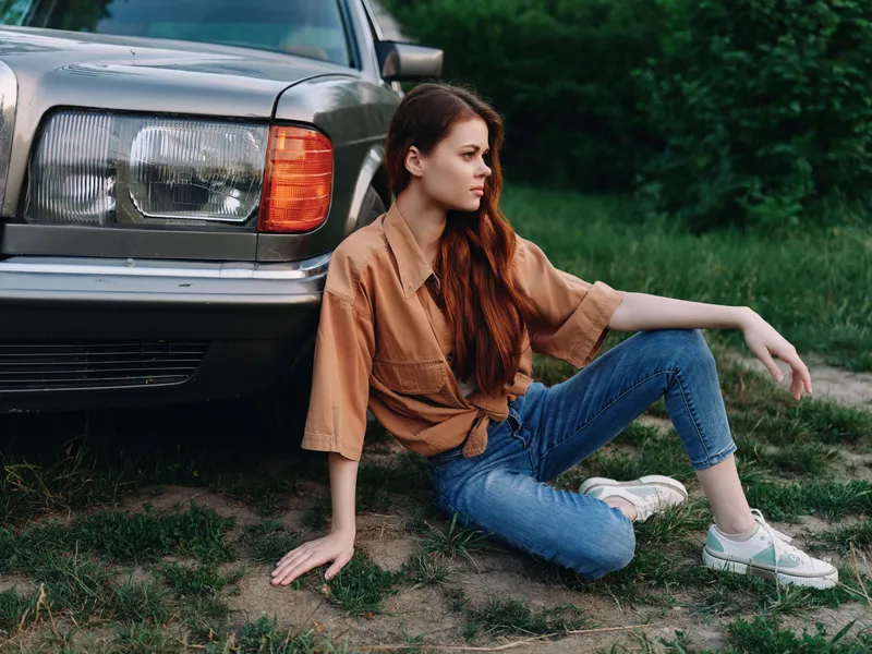 Young woman in casual clothes sitting next to vintage car in green environment, showcasing a relaxed summer vibe, with a focus on fashion and freedom.