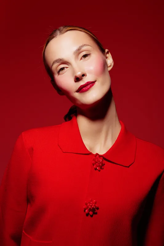 Portrait of a confident woman in red sweater with decorative buttons against red background. Studio lighting highlights her clear skin and subtle smile. Concept of fashion and elegance.