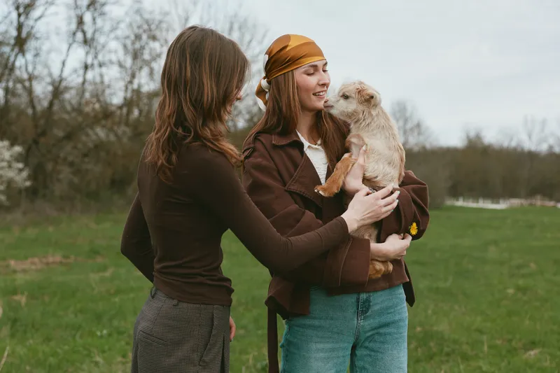 Outdoor lifestyle scene with two women smiling as one holds a goat kid, softly lit by natural light, casual jackets, grassy meadow background, carefree connection, warm tones, candid moment.