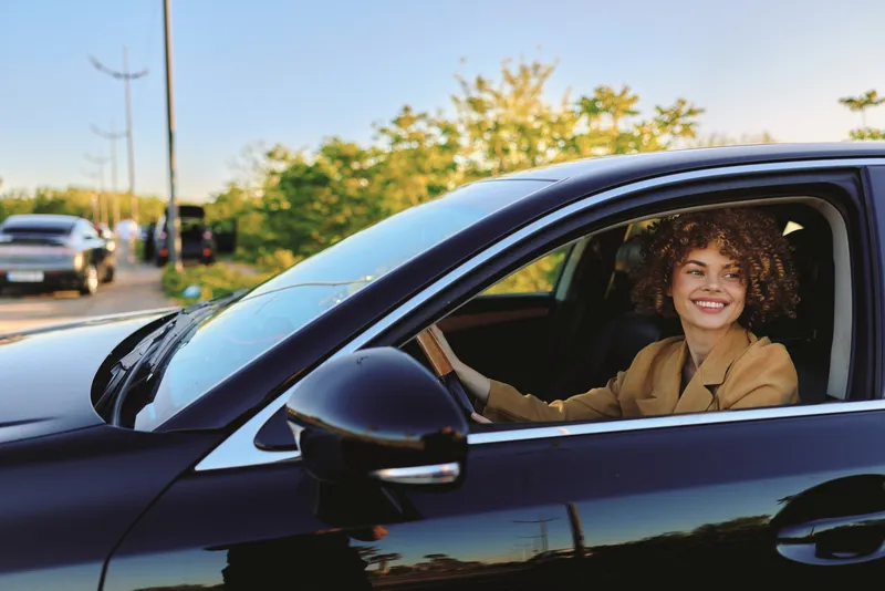Driving young woman with curly hair sits in a sleek car with a bright smile, enjoying a carefree moment in retro film color outdoors
