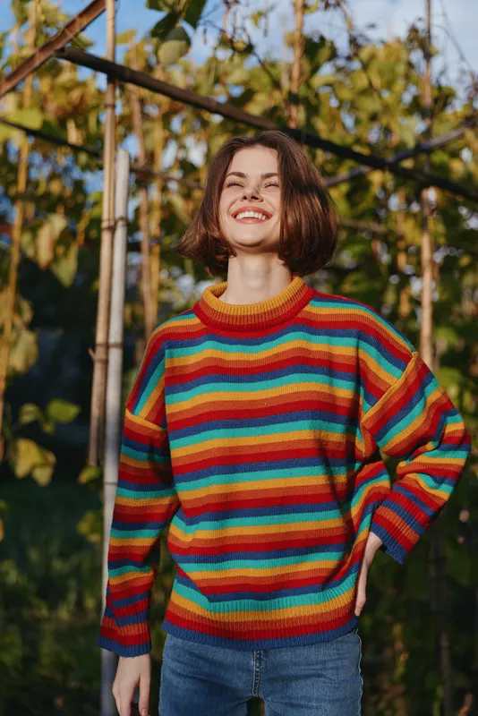 Young adult woman smiling in a colorful striped sweater with short hair, casual outfit, leaning with hand in pocket in an outdoor garden among sunlit vines, joyful relaxed lifestyle portrait.