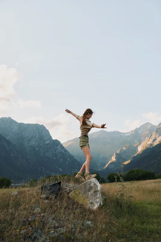 Young woman balancing on a large rock in a mountain meadow during daytime. Outdoor lifestyle concept with nature, adventure, and freedom in a scenic landscape.