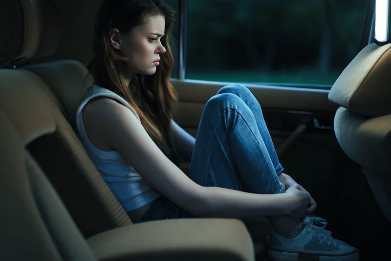 Young woman sitting alone in a car, looking pensive and sad, wearing casual jeans and a tank top, reflecting on her emotions in a darkened environment.