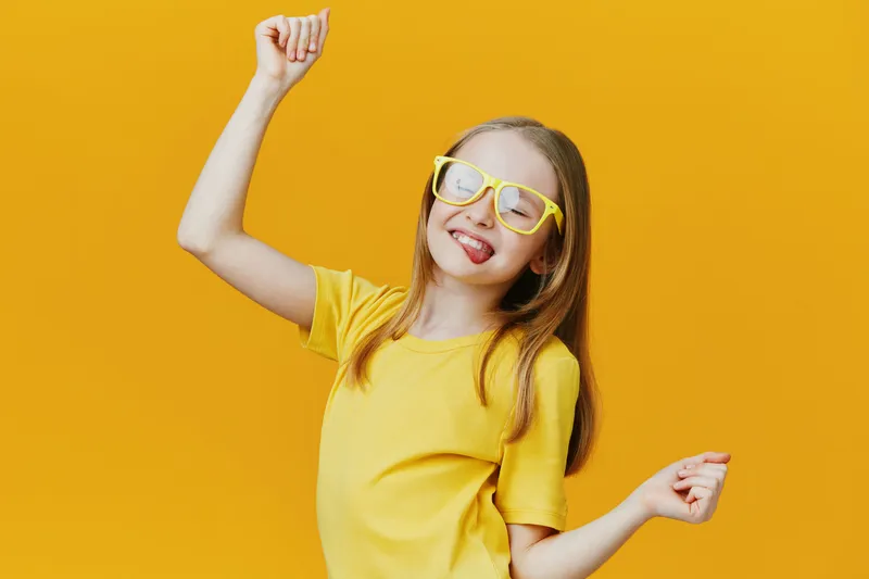 Happy girl with yellow glasses dancing against a vibrant yellow background, expressing joy and confidence in a casual outfit, perfect for cheerful concepts