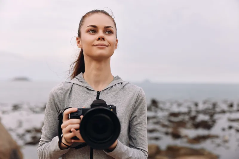 Confident young woman holding a camera by the beach, wearing a gray hoodie, against a serene ocean background, showcasing outdoor photography passion