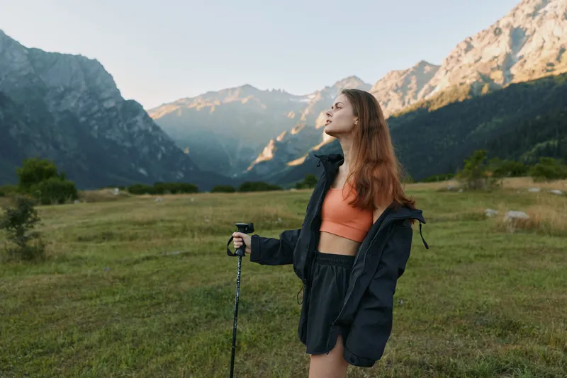 A cheerful hiker stands in a scenic mountain meadow, looking upward with a trekking pole in hand. She wears an orange top and a dark jacket, embodying adventure and outdoor energy.