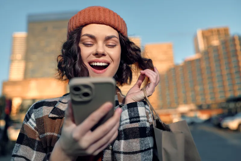 Woman smartphone shopping smile city beanie bag sunlight in urban street at golden hour, plaid coat candid lifestyle portrait with authenticity, mindful living and emotional storytelling.