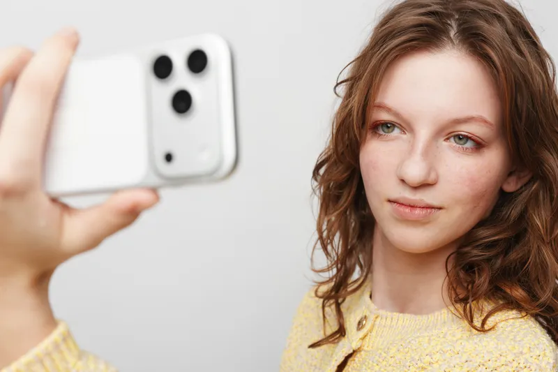 Young woman with curly hair wearing yellow sweater takes selfie with smartphone on white background. Concept of social media, technology, and casual lifestyle portrait.
