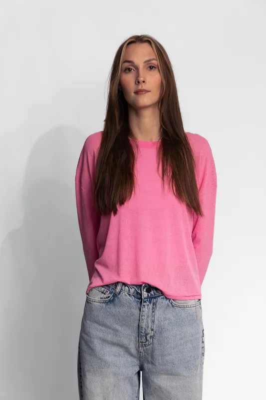 Young woman with long brown hair wearing a pink sweater and light blue jeans standing against a plain light background. Casual fashion and confident pose concept.